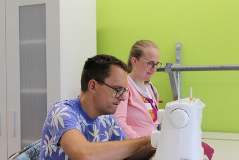 Ein Mann sitzt konzentriert an einer Nähmaschine und näht. Eine Frau im Hintergrund schaut zu.